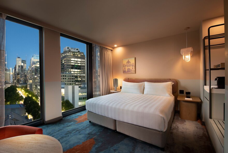 A glowing hotel room at dusk with a king size bed and a city view through the windows.