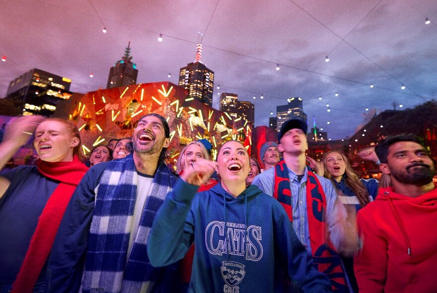 Football fans, some smiling and others frowning, at dusk at Fed Square with lights and buildings in the background.