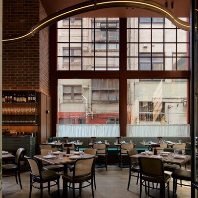 The double height interior of a restaurant with tables and chairs set for service in front of a large window. 