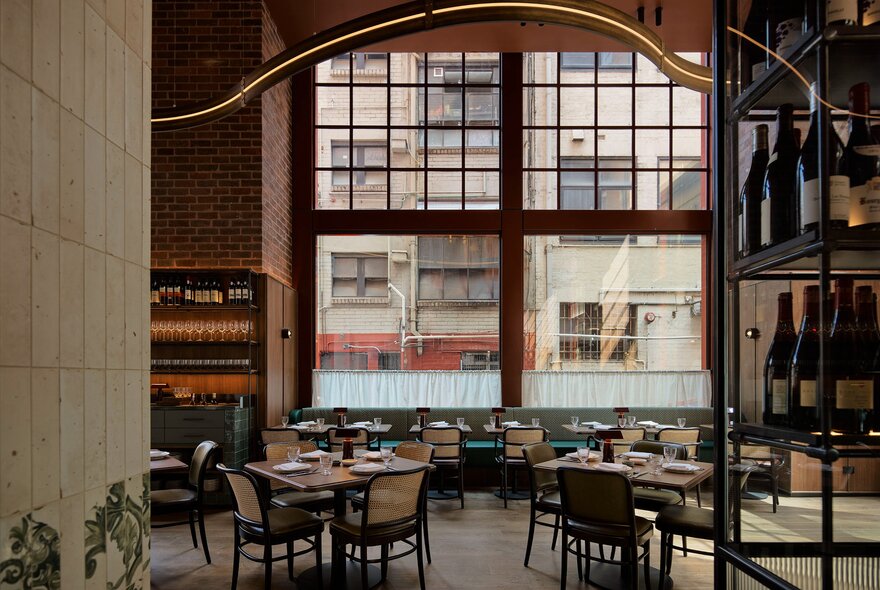 The double height interior of a restaurant with tables and chairs set for service in front of a large window. 