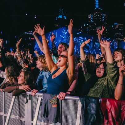 A live concert event at Fed Square at night, with a large, enthusiastic crowd gathered near a stage barrier.