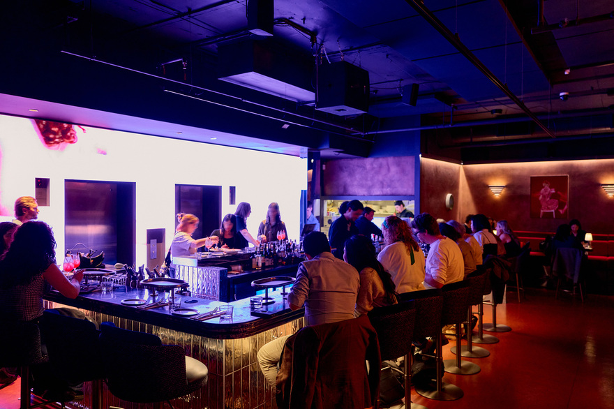 A busy restaurant with people dining and drinking at a bar surrounded by purple neon lights