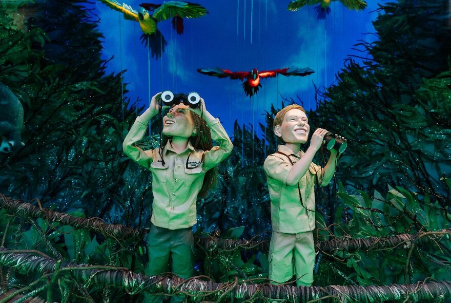 The Myer Christmas Window display, showing two explorer figures wearing khaki clothes, holding binoculars, in the Australian rainforest with colourful tropical birds flying above them.