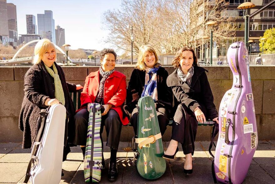 Four female classical musicians holding instrument cases, seated on a bench on a bridge near the Yarra with city views in the background.