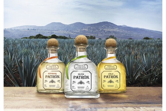 Three tequila bottles resting on a wooden table in front of an agave plantation, mountains in the distance.