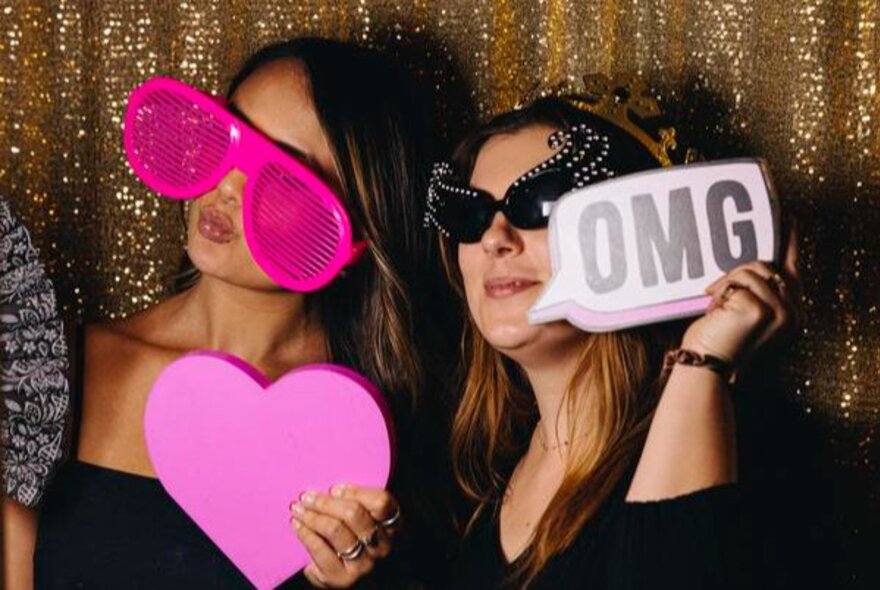 Two women, one with giant pink novelty glasses holding a pink heart, the other holding speech bubble that says OMG.