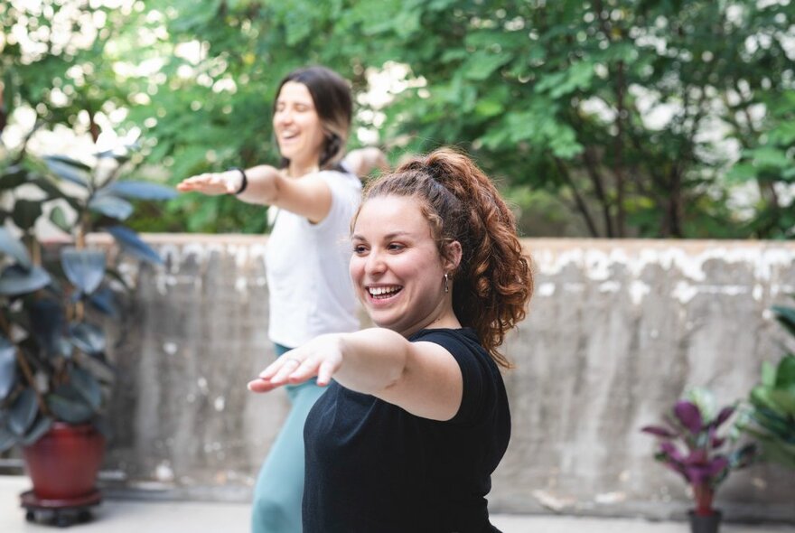 Two women smiling and doing pilates outdoors in a leafy space. 