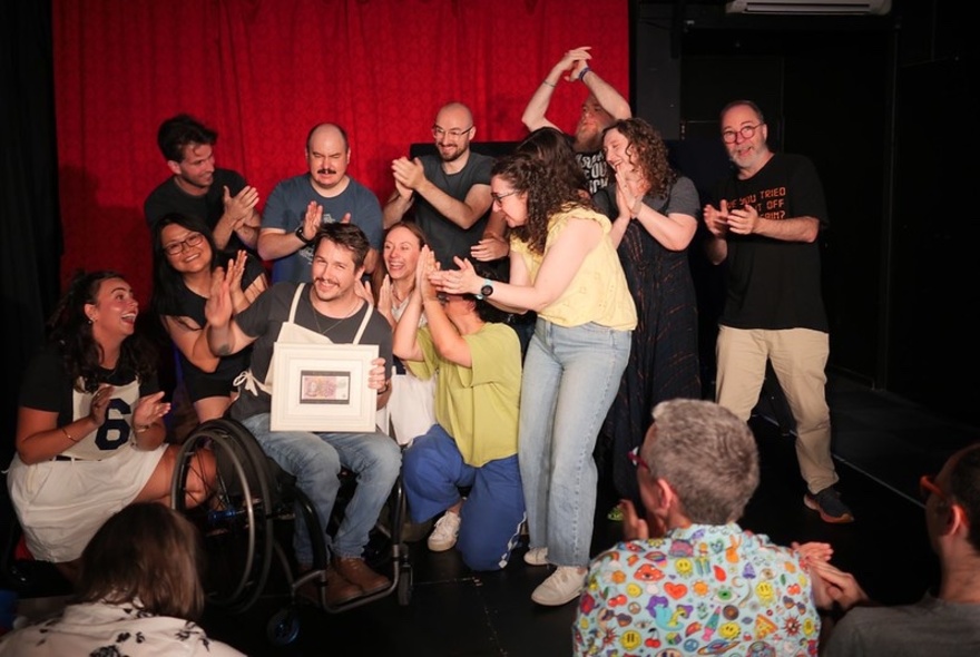 An ensemble cast of impro actors on a small theatre stage, all congratulating a person who is holding up a certificate.
