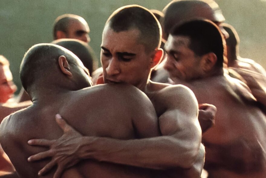 A still image from a film showing men naken from the waist up embracing in soft light. 