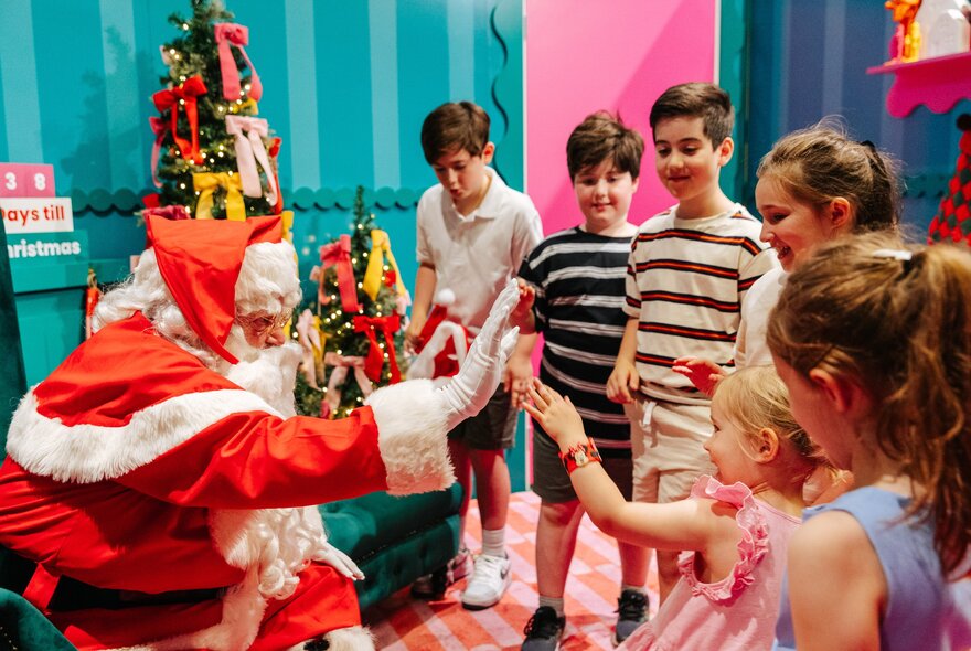 Santa surrounded by a group of young children, holding his hand out to high-five a little girl in pink.