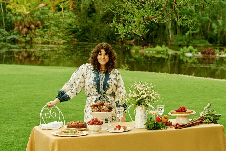 Chef Julia Busuttil Nishimura standing at a table set with dishes and flowers in the Botanic Gardens, her hand on a decorative garden seat.