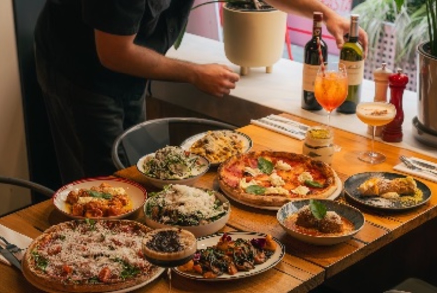 Hands arranging bottles next to a wooden table laid with manyy dishes of food including pizzas.