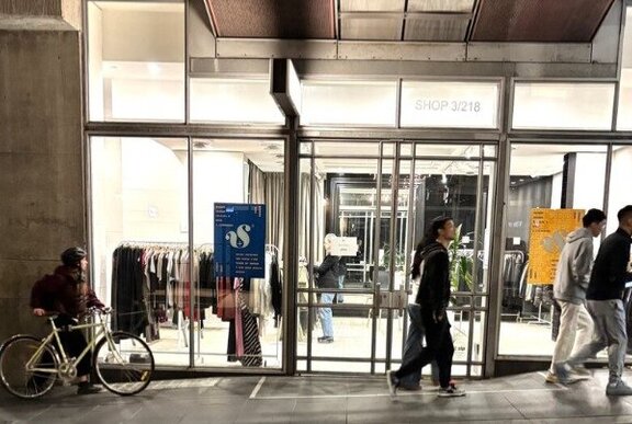People walking past a brightly lit shop front with clothes on display behind the glass windows and doors.