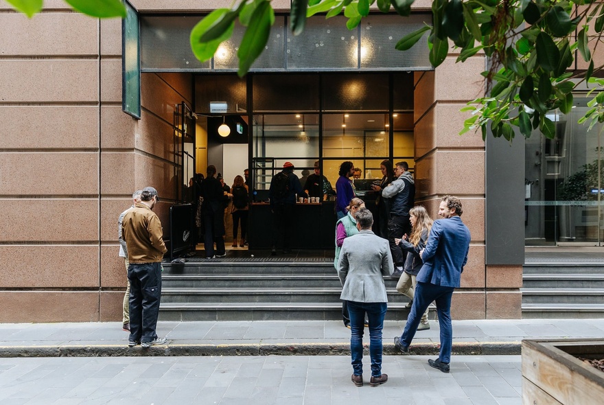 People gathering outside a city cafe in a business-like stone building with steps. 