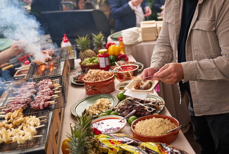 A person selecting food from a open buffet table and grill area, and placing it onto a plate they hold in their hands.