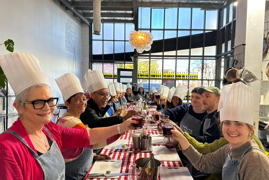 People seated around a long table in a restaurant, all wearing chef hats and smiling as they hold up cups of their hand-made sangria.