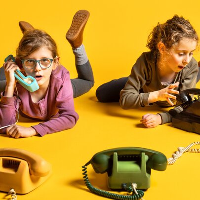 Two girls on their tummies on a yellow floor talking on landline telephones. 