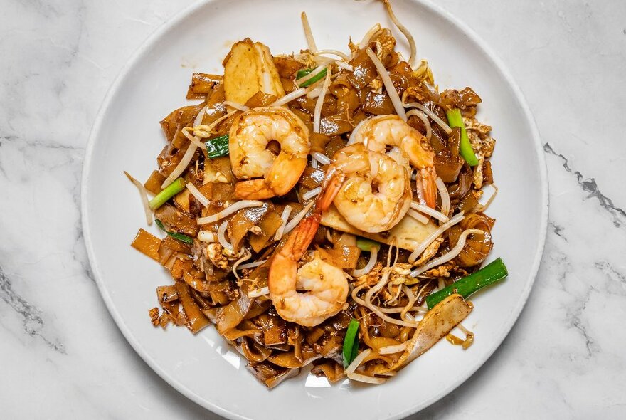 Looking down at a white dish of noodles, prawns and vegetables, on a table.