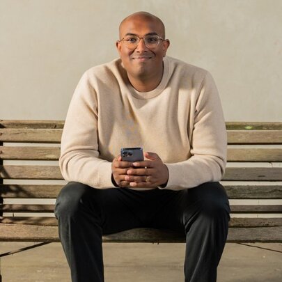 Comedian Rowan Thanbar sitting on a park bench, inside, smiling and holding his phone. 