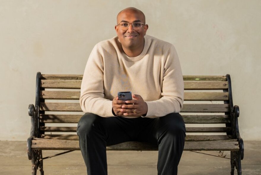 Comedian Rowan Thanbar sitting on a park bench, inside, smiling and holding his phone. 