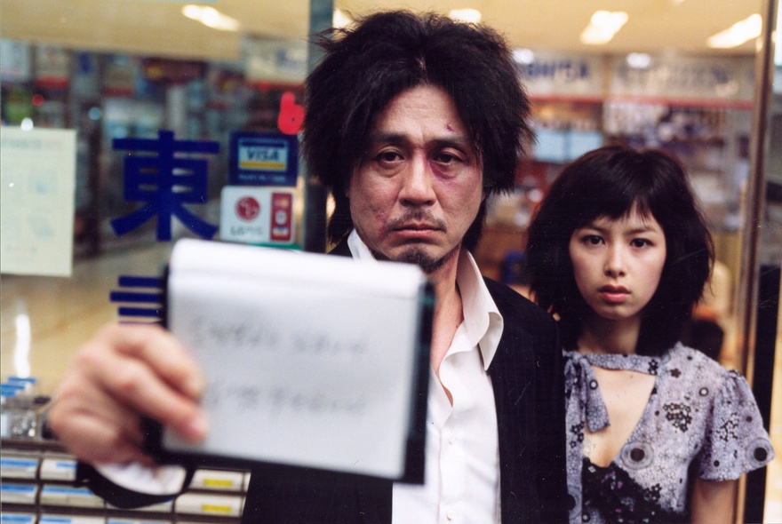 A still image from the film Oldboy with a sad man holding forth a handwritten note, a woman beside him looking blank.