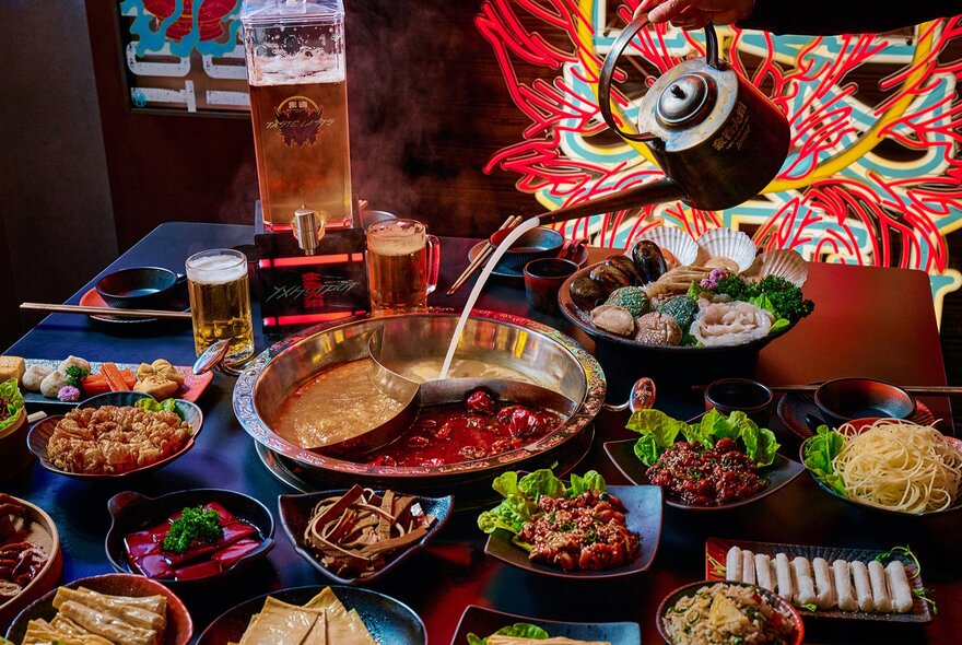 A teapot pouring soup into a sectioned hotpot surrounded by colourful side dishes.