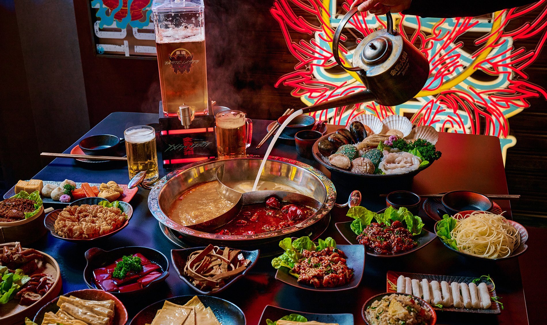 A teapot pouring soup into a sectioned hotpot surrounded by colourful side dishes.