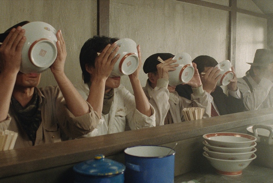 A line of Japanese men seated at a bar in a ramen restaurant in Japan, each man drinking the soup from the bowl.