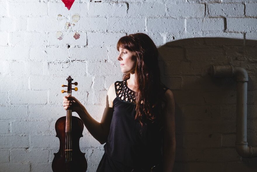 A woman in holding a violin up to her right, looking down at it, in a room with a painted brick wall. 