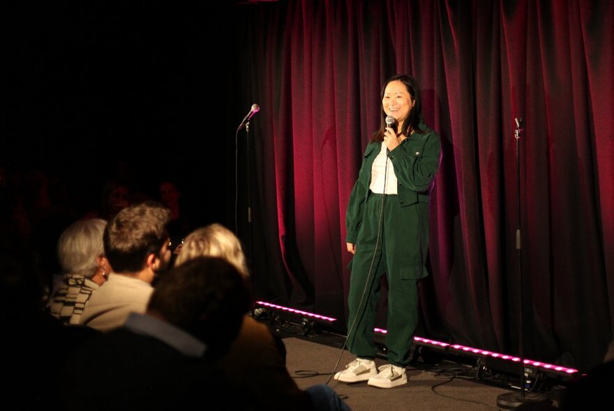 A comedian standing on a stage and looking relaxed in front of an audience, holding a microphone and performing stand-up comedy.