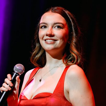 A singer in a red dress, smiling and holding a microphone while on stage, a purple stage curtain behind her.