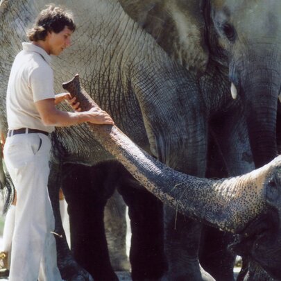 A man in 70s clothes holding the trunk of an elephant, another elephant and man behind. 