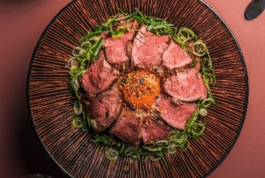 Looking down on a plate of Steak Don, with an egg yolk in the middle of arranged rare steak slices. 