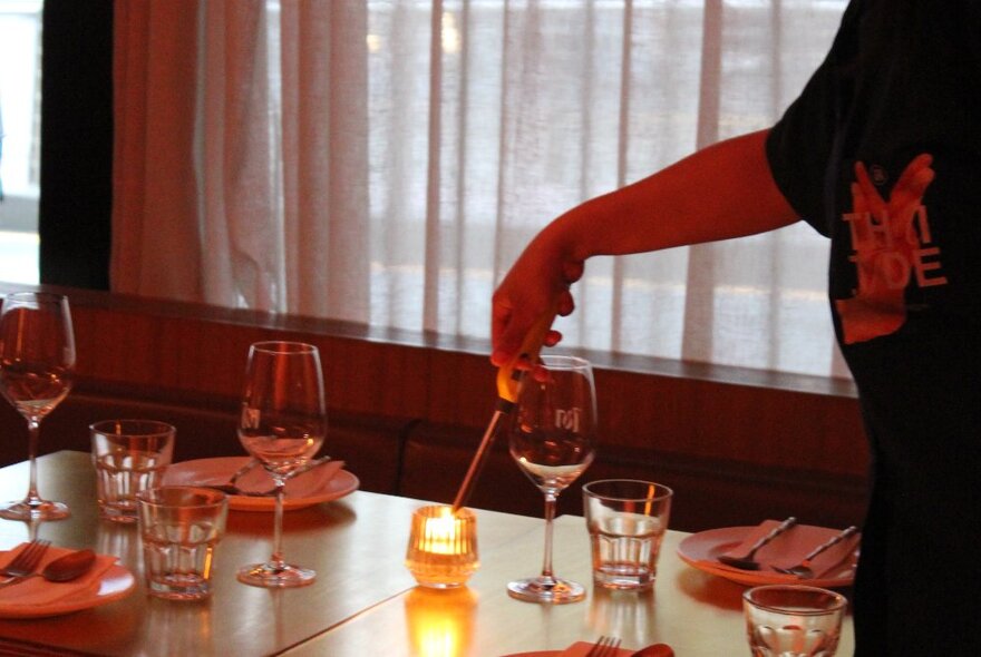 A waiter lighting a candle on a table set for dinner, sheer curtains over a window behind.