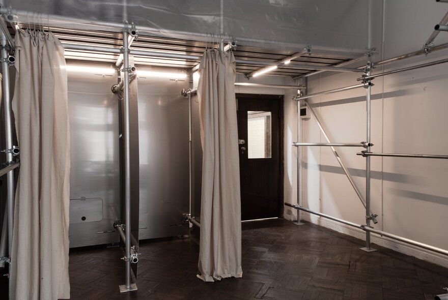 A bare, colourless looking space with a door, metal scaffolding bars and two change rooms with curtains. 