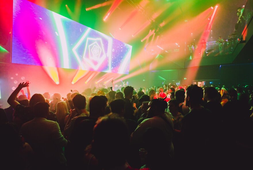 Crowd of people standing under strobe lights in front of a large screen and stage.
