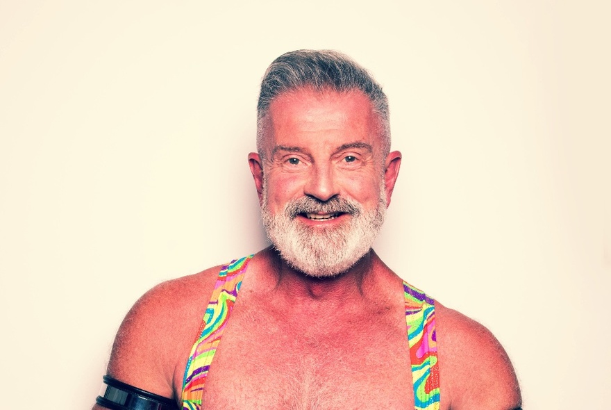 A portrait of comedian, Brent Thorpe, from the chest up wearing brightly coloured straps over his shoulders, against a cream background.