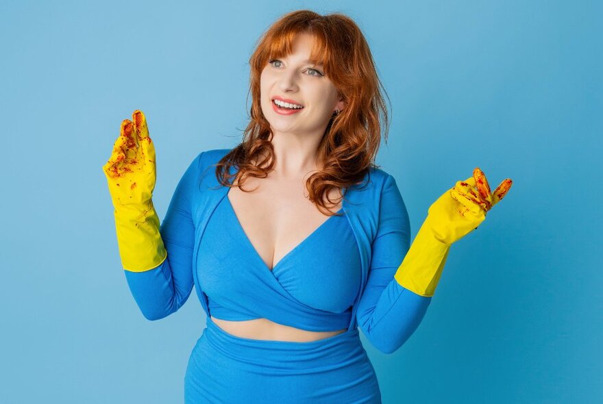 Performer Robyn Reynolds, a woman wearing an aqua coloured tight fitting dress, yellow rubbers gloves stained red at the fingertips, with shoulder length red hair and with a smile on her face.