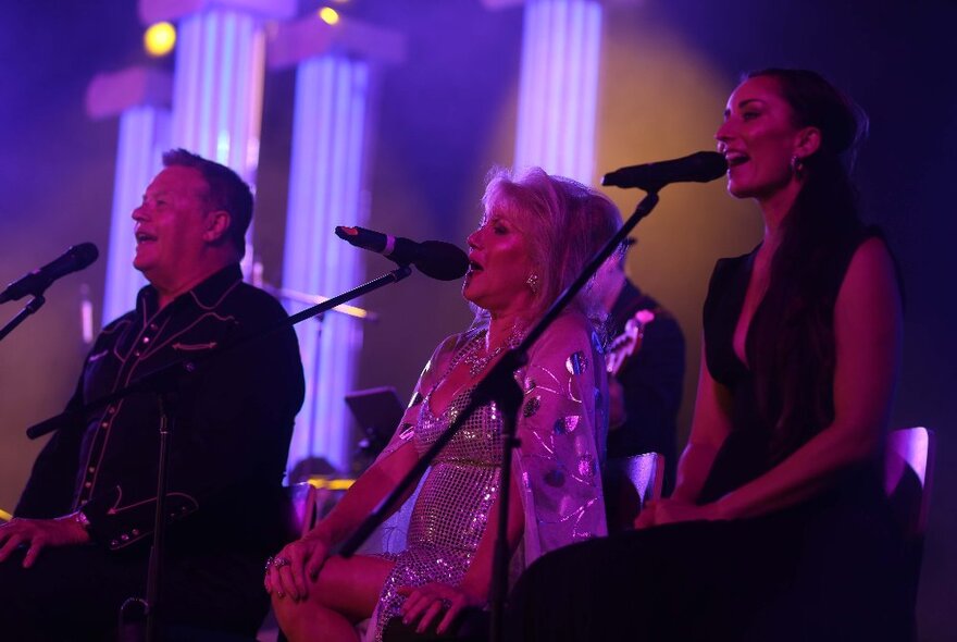 Three singers singing into microphones on stage at night, with purple-lit pillars behind them.