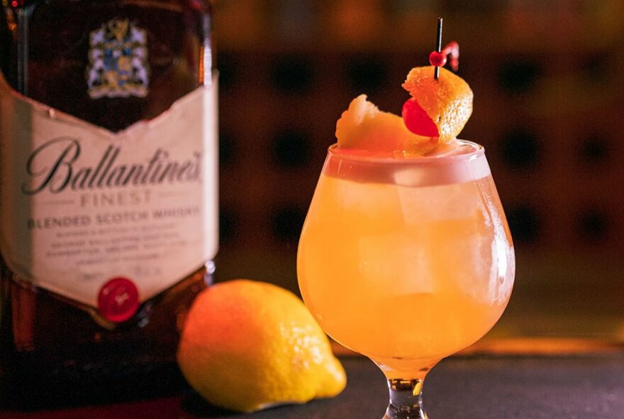 An orange coloured cocktail in a bell glass, with a citrus peel garnish and a bottle in the background.