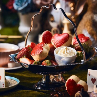 A high tea spread with heart-shaped scones in a silver basket, cups of tea and playing cards on the table. 