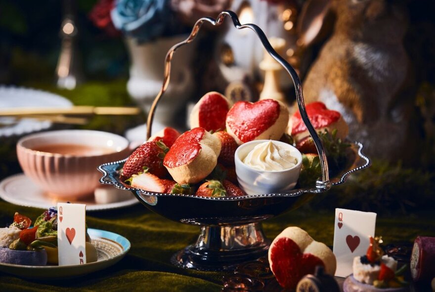 A high tea spread with heart-shaped scones in a silver basket, cups of tea and playing cards on the table. 