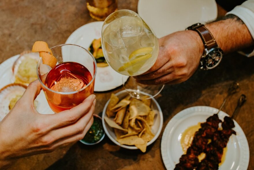 Two hands clinking filled glasses over dishes of food, on a restaurant dining table.