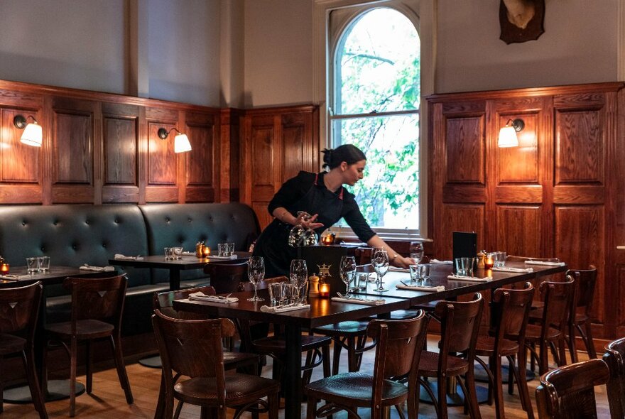 Dining room of a restaurant with a long table being set with glasses and tableware by a waitress, leather banquette seating, wood panelled walls and soft lamp lighting.