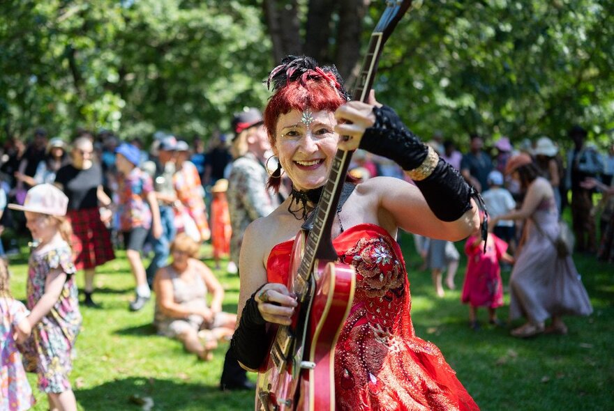 A musician playing a red guitar, wearing a strapless red dress with red hair, children and picnickers in the background under trees.