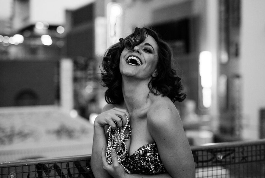 A black and white image of a woman in a strapless gown throwing her head back and laughing while holding pearls in her hand. 