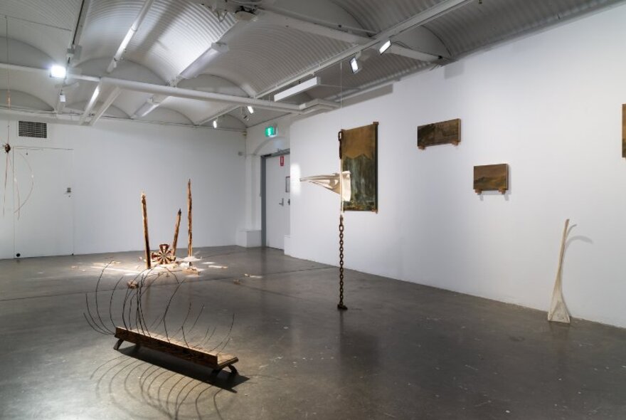 A sparse gallery space with white walls and a concrete floor, with wire sculptures and framed wall art displayed.