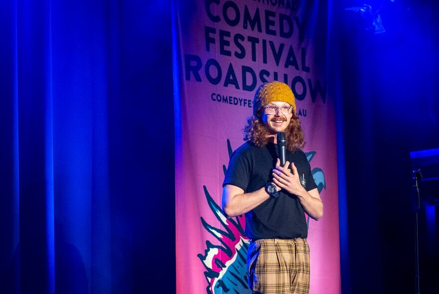 Comedian with curly red hair and glasses wearing an orange beanie, standing on stage holding a microphone.