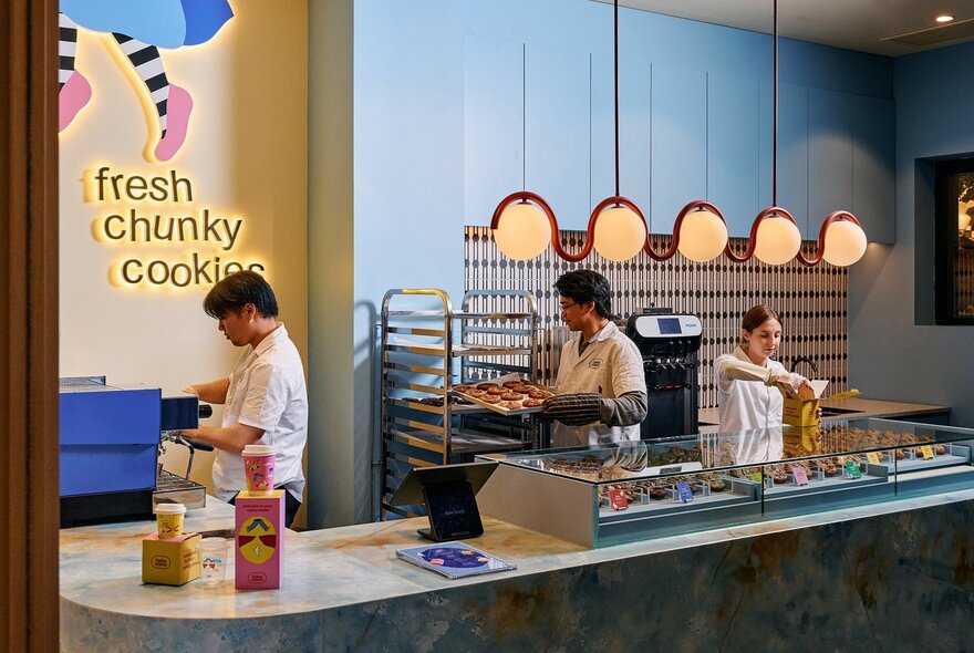 The interior of BakeStand, a small bakery, with a central counter, pendant lighting, pale blue walls and three staff at work.