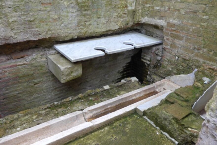 Ancient Roman public toilet showing a bench seat with cut out holes, positioned above a sewer trough, all contained in a walled brick structure.